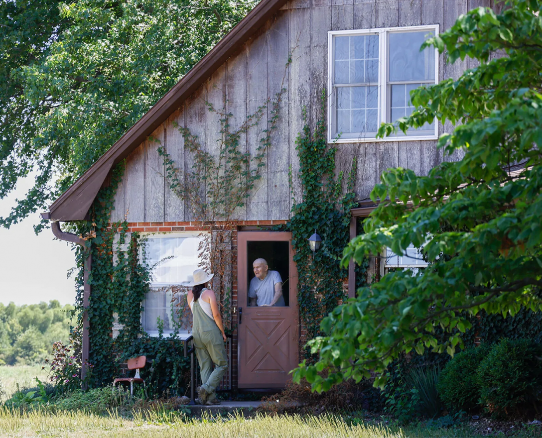 A man and a woman stand at the doorway of a wooden farmhouse. Lush green trees frame both sides of the house. 